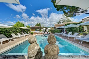 Une piscine entourée de verdure et de bâtiments, avec des chaises longues et des parasols.