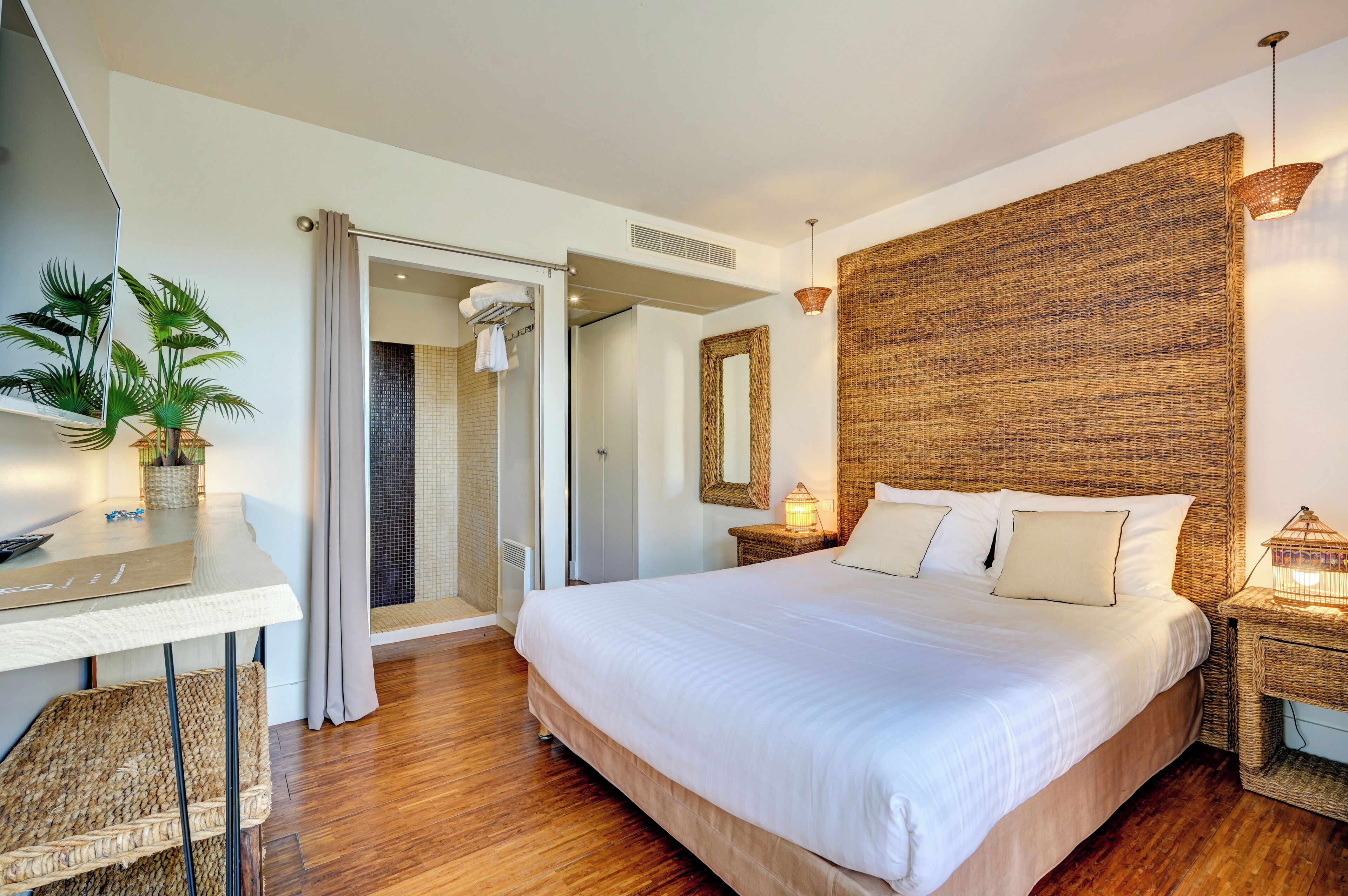 A modern hotel room with a large bed, a desk, a shower area, and a wicker headboard.