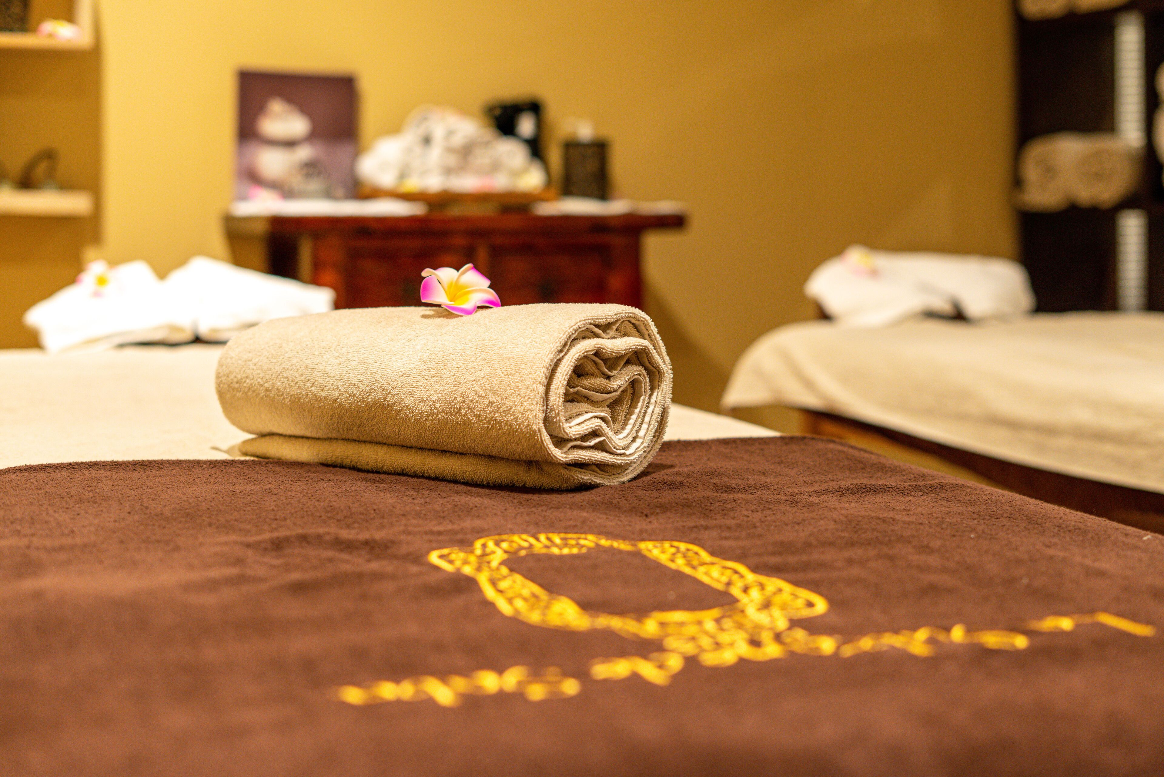 A neatly made bed with a rolled towel and a decorative flower, a brown towel with a golden design, and a wooden nightstand with a framed picture and a bottle in the background.