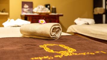 A neatly made bed with a rolled towel and a decorative flower, a brown towel with a golden design, and a wooden nightstand with a framed picture and a bottle in the background.