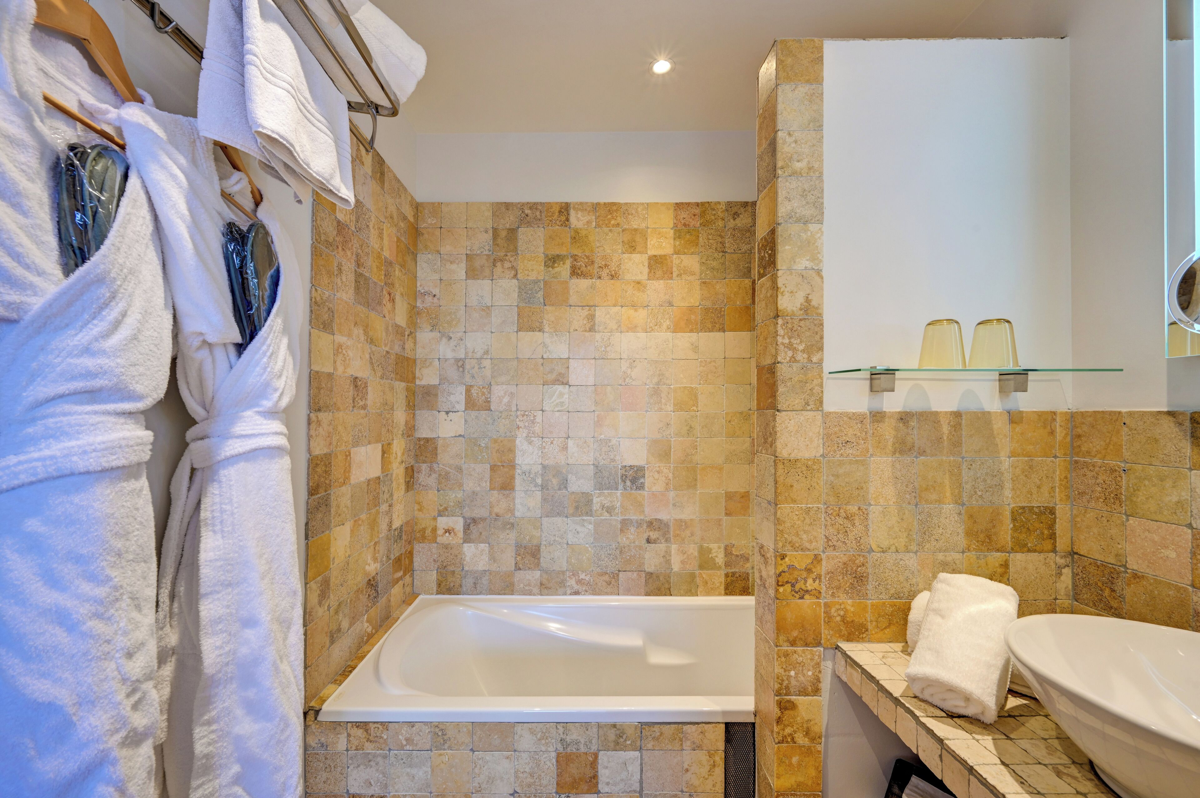 Une salle de bain avec une baignoire, des murs carrelés, une étagère en verre où sont posés les articles de toilette, et des serviettes suspendues à un porte-serviettes.