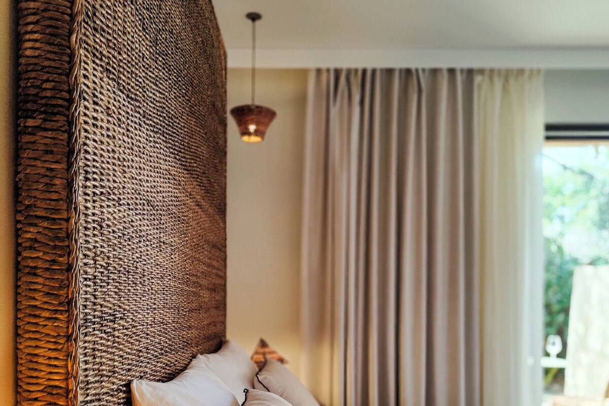 Una cama bien hecha con almohadas, un cabecero tejido, una lámpara colgante y vistas a la vegetación exterior.