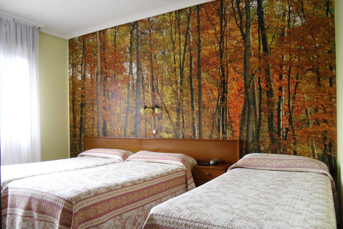 A hotel room with two beds, a large forest mural on the wall, and a nightstand with a lamp.