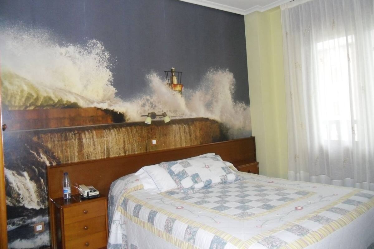 A hotel room with a large wall mural of a crashing wave, a wooden headboard, a bed with a floral quilt, a nightstand with a bottle and a phone, and a window with sheer curtains.