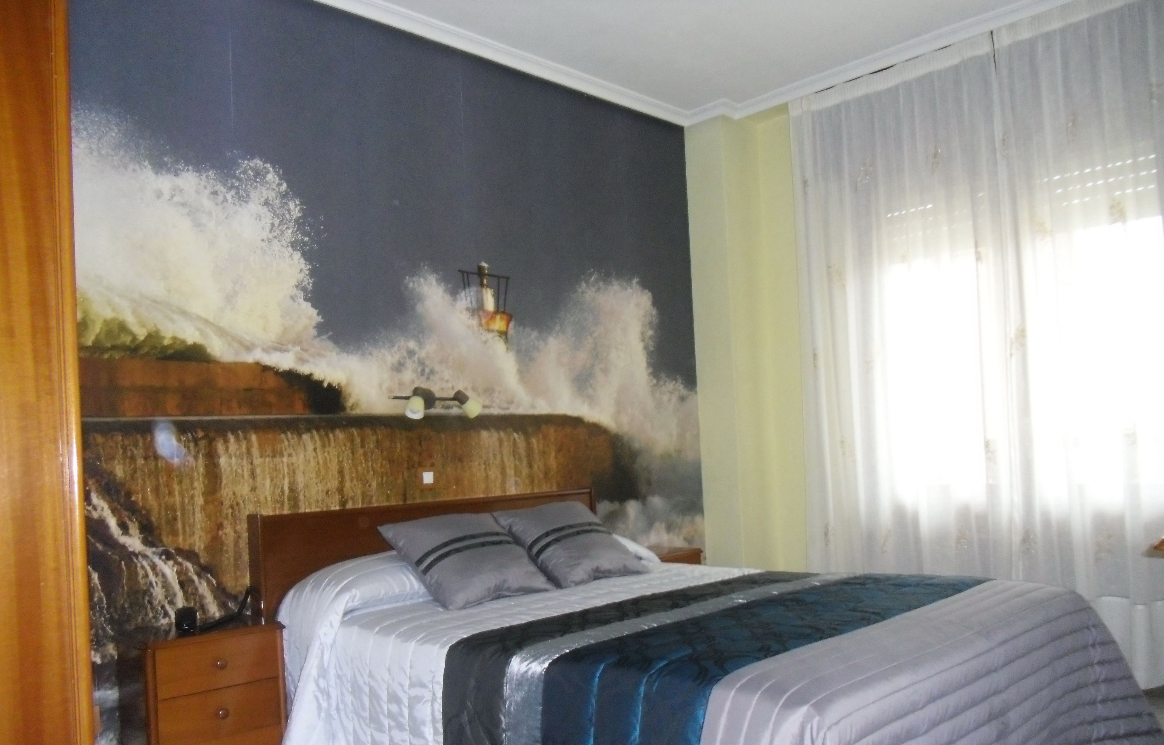 A bedroom with a large bed, a nightstand, a wall-mounted lamp, and a window with sheer curtains.