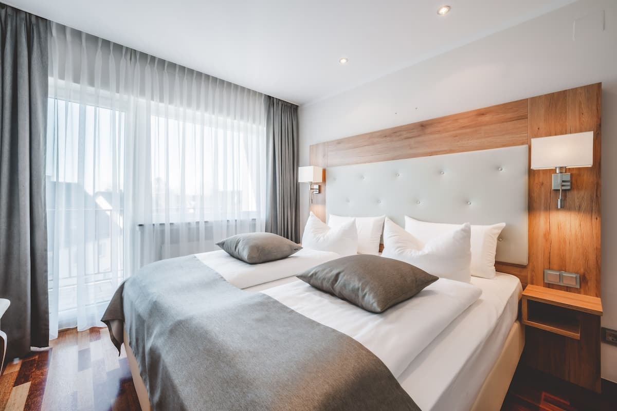 Ein modernes Hotelzimmer mit einem großen Bett, zwei Nachttischen, einem Holz-Kopfteil und einem großen Fenster mit Vorhängen.