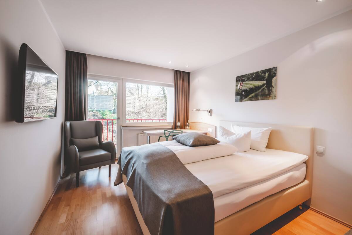 Ein modernes Hotelzimmer mit einem großen Bett, einem Sessel, einem Schreibtisch und einem Fenster mit Blick ins Freie.