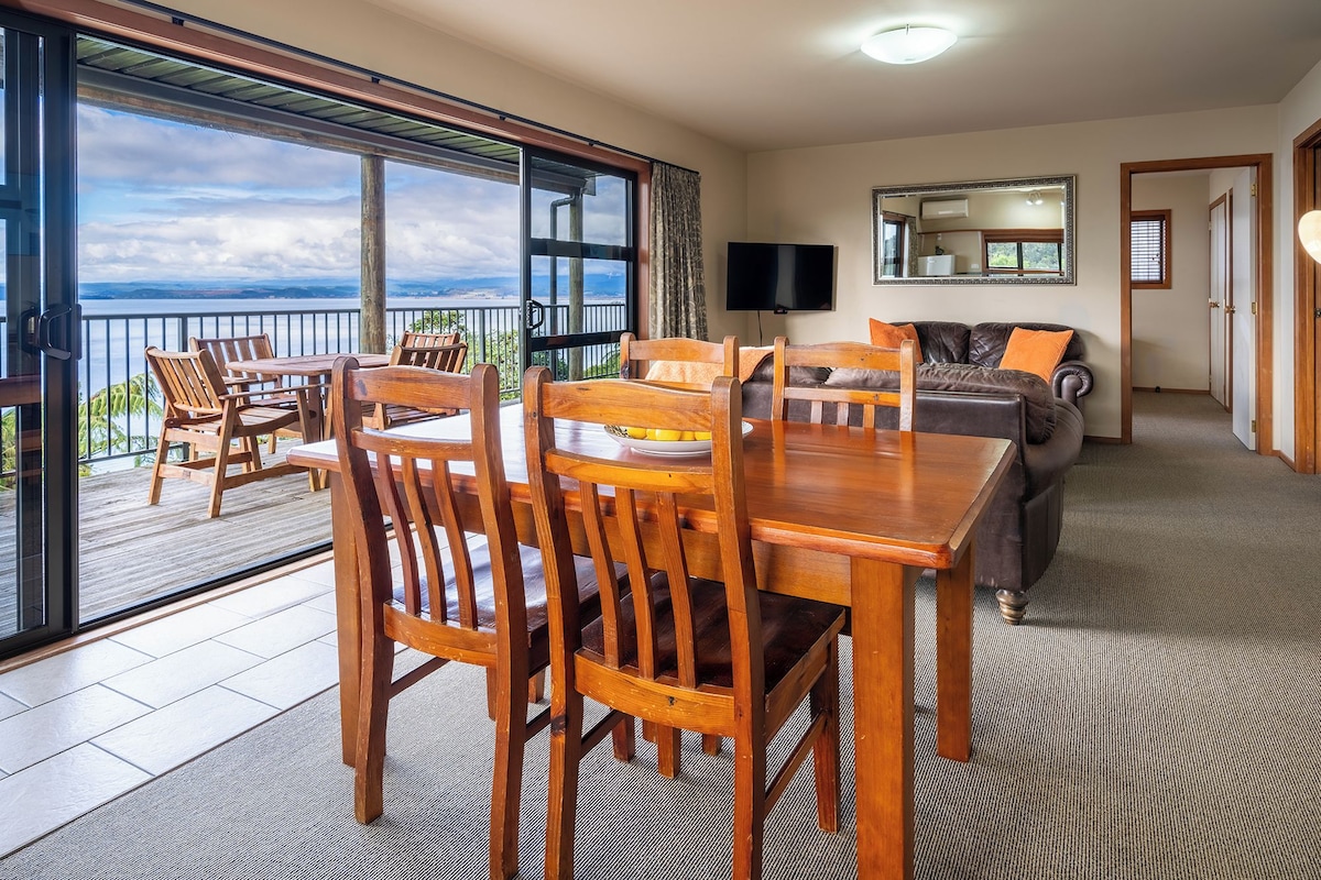 A spacious living area with a wooden dining table, chairs, a sofa, and a balcony with a view.