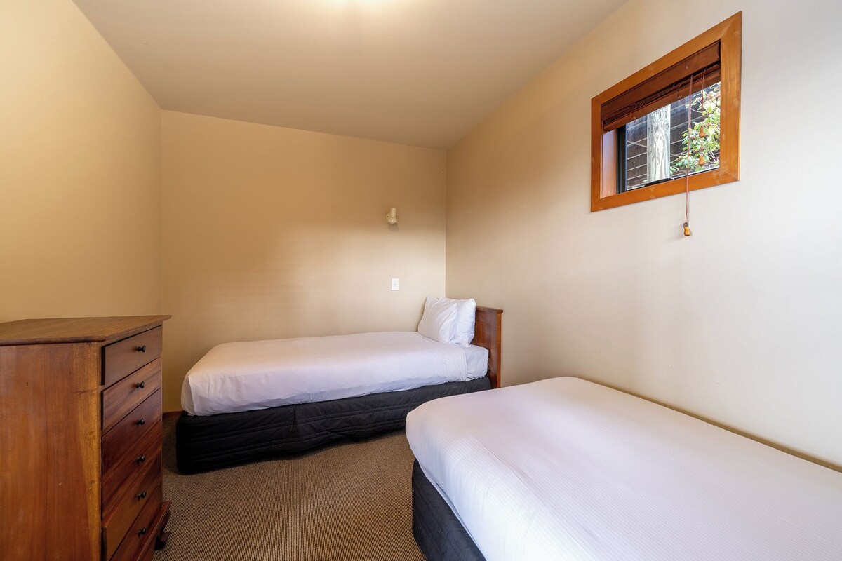 A small, single-room accommodation with two single beds, a wooden dresser, and a window with a view of trees.