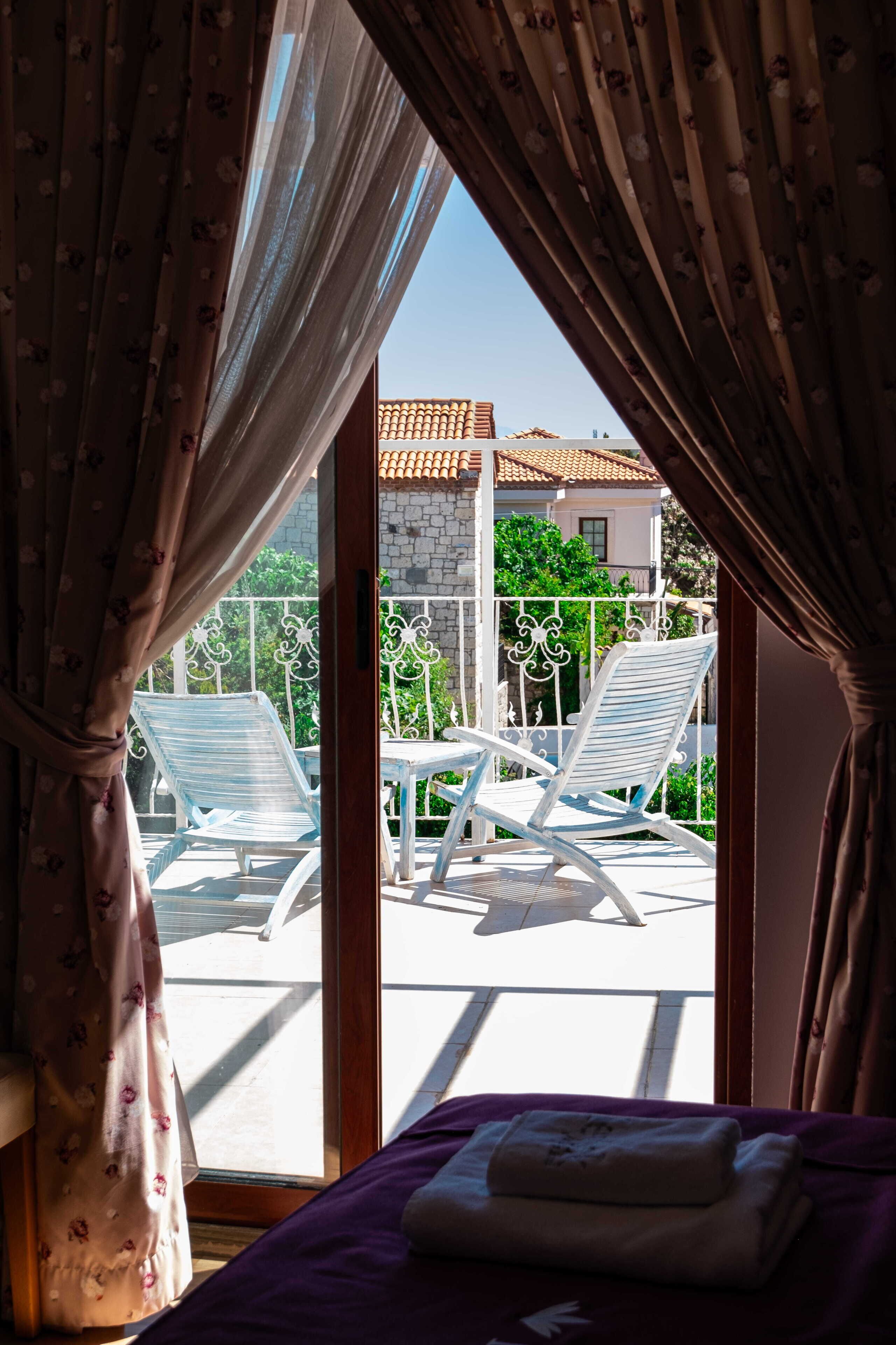 Una habitación con cama, ventana con cortinas y vista a un balcón con dos sillas blancas y una mesa.