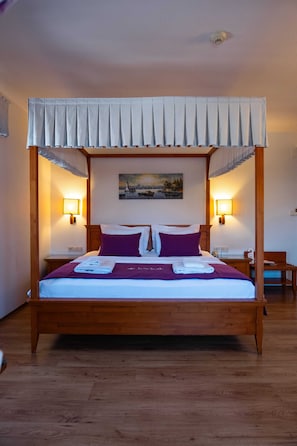 Uma cama de dossel com cortina, duas luminárias de cabeceira, um quadro na parede e um criado-mudo de madeira com um telefone.