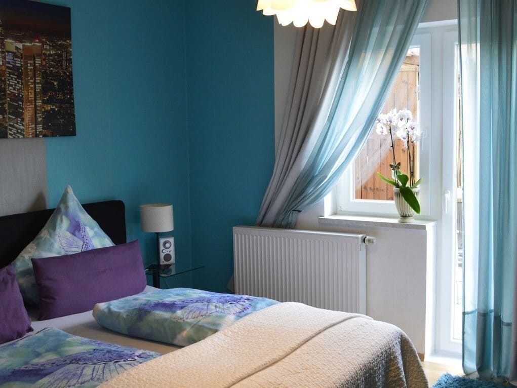 A bedroom with a bed, pillows, a nightstand, a lamp, a window with curtains, and a cityscape painting.