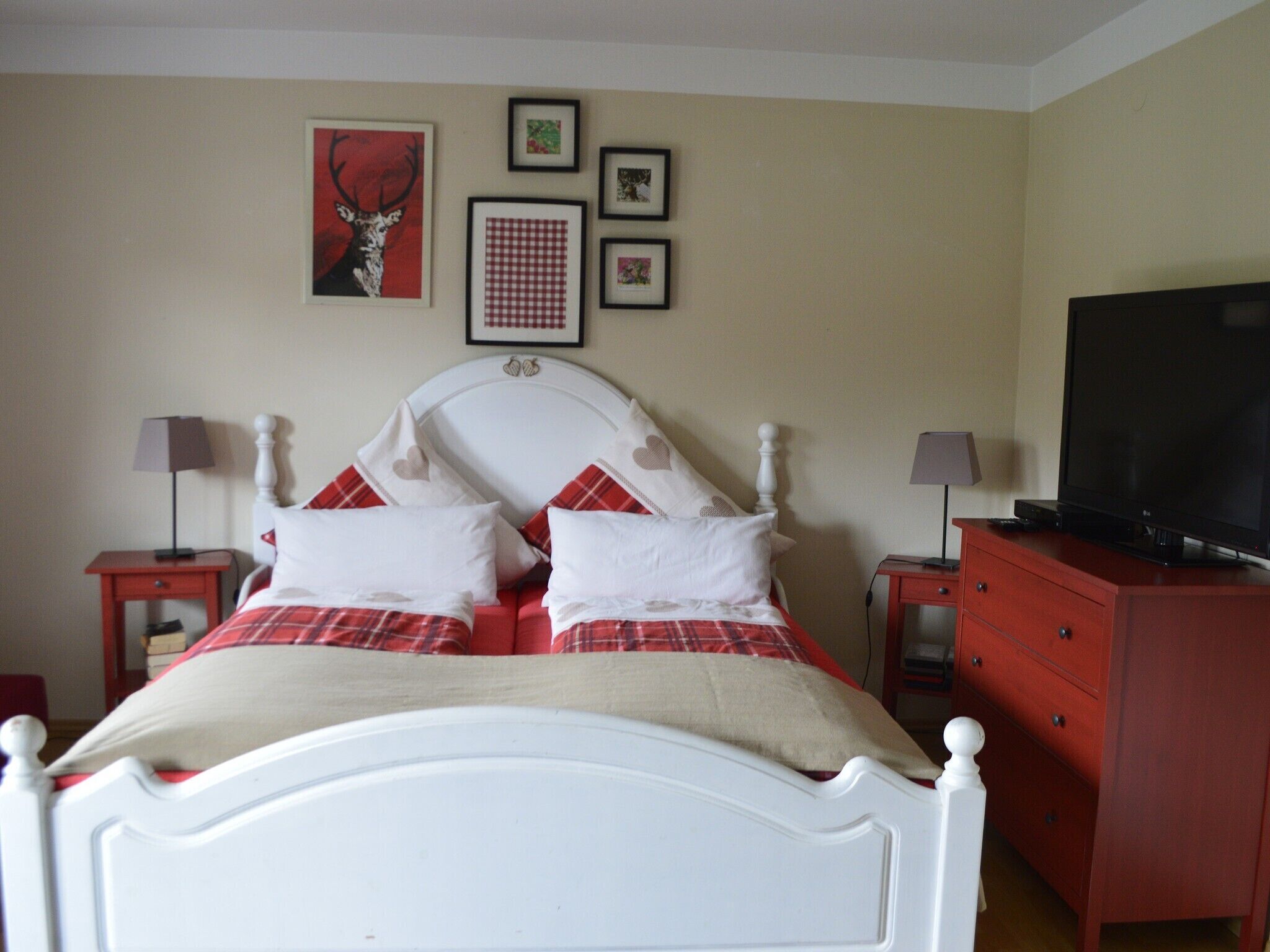 A bedroom with a white bed, red and white bedding, a television on a dresser, and framed pictures on the wall.