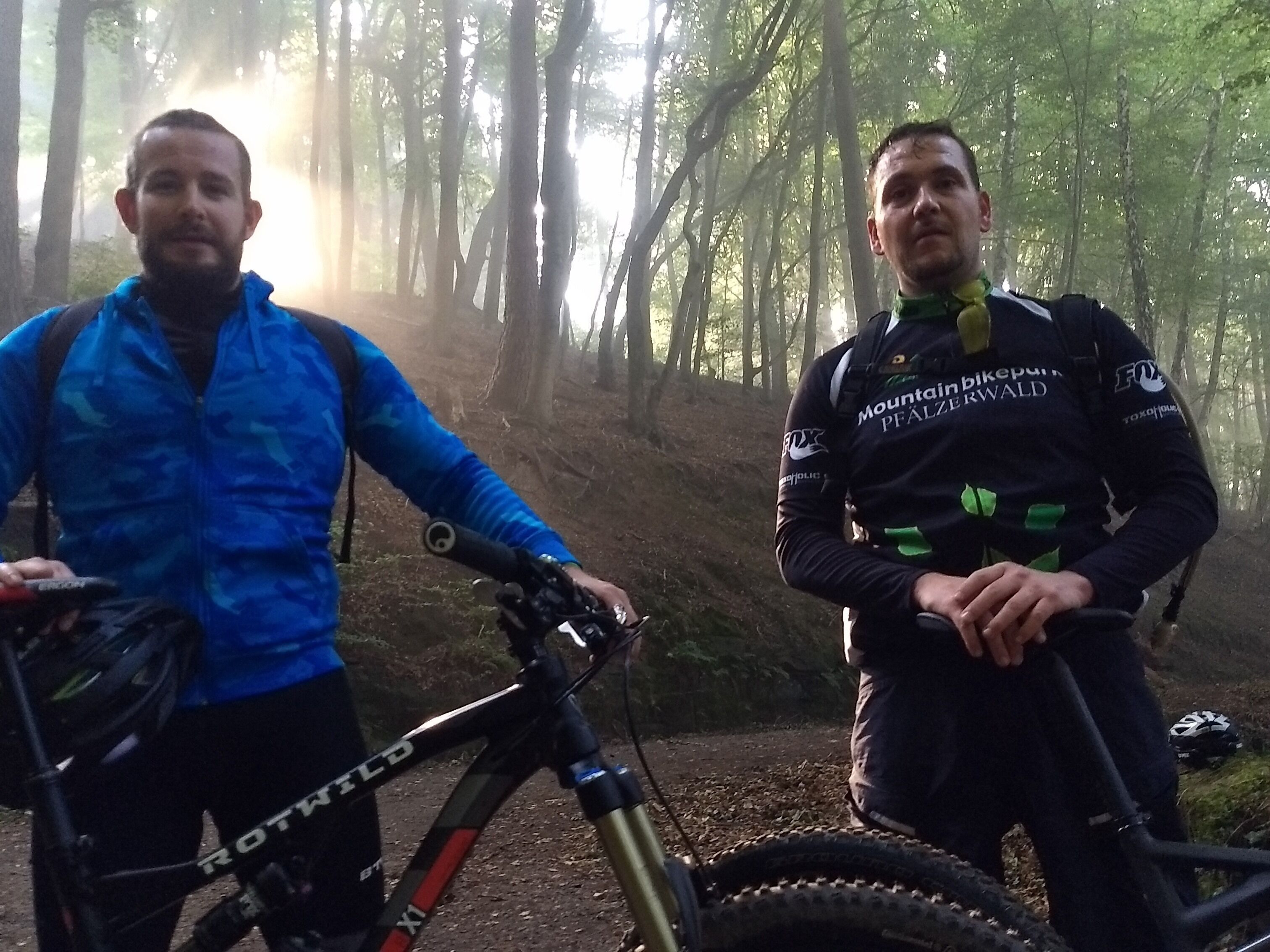 Zwei Personen stehen neben Mountainbikes in einem bewaldeten Gebiet.