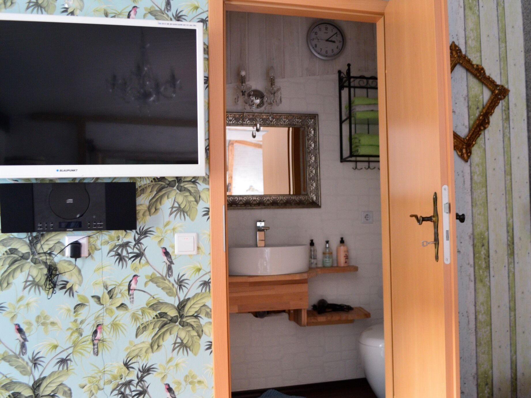 Une salle de bain avec un meuble-lavabo en bois, un miroir, une horloge et un téléviseur fixé au mur.