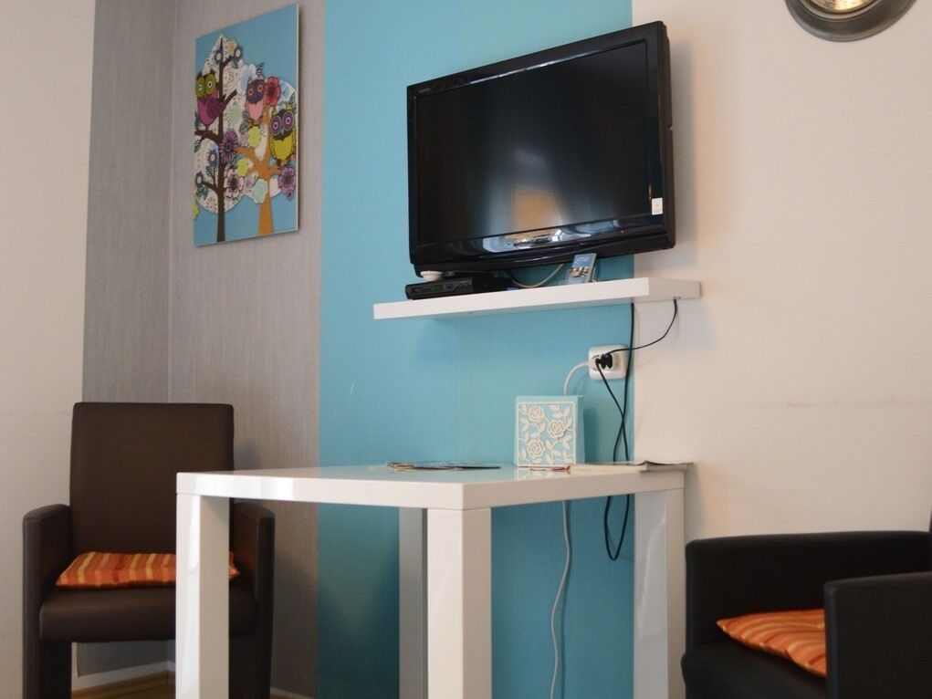 A living room with a flat-screen TV mounted on a white shelf, a small table with a cup, and two chairs with orange cushions.