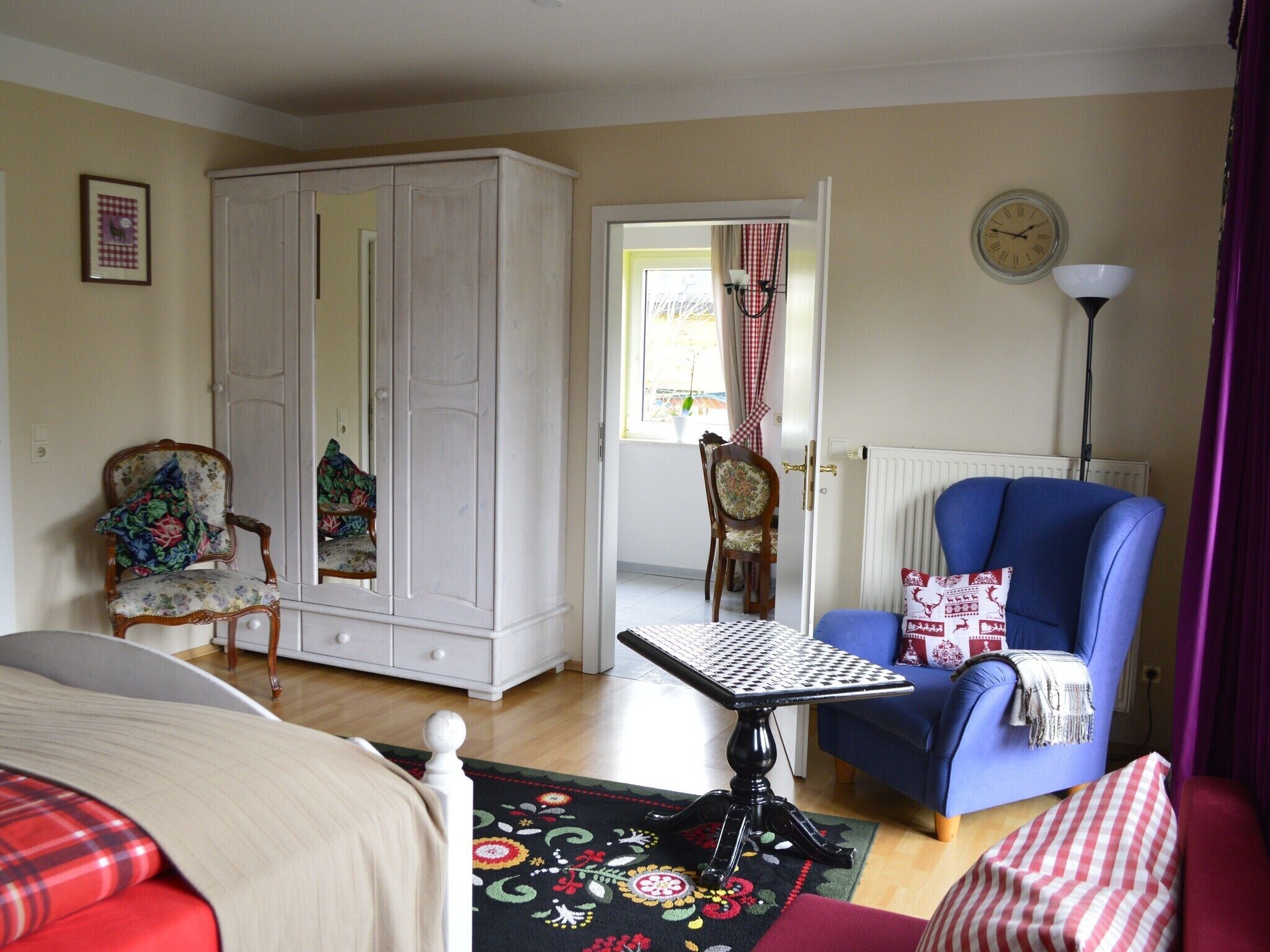 Una habitación con una cama, un sillón azul, un armario blanco, una silla de madera, una mesa con mantel de cuadros y una lámpara de pie.