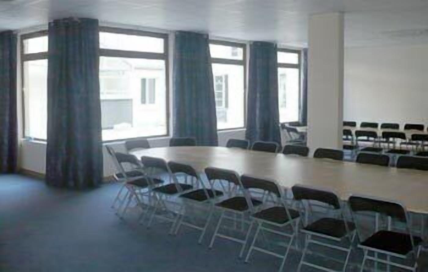 Un salón de conferencias con una amplia mesa ovalada rodeada de sillas, ventanas grandes con cortinas y una pared blanca lisa.