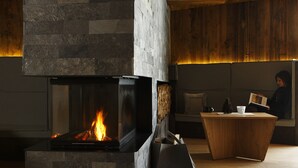 Une cheminée moderne avec une façade en pierre, un banc en bois et, au fond, une personne qui lit un livre.