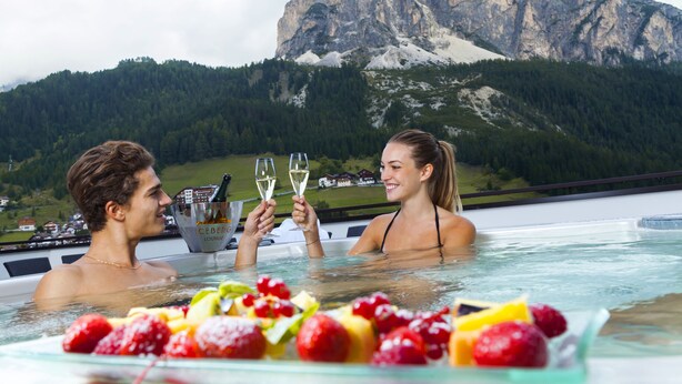 Un couple dans un bain à remous, avec une vue sur les montagnes, tenant des coupes de champagne, et un panier de fruits au premier plan.