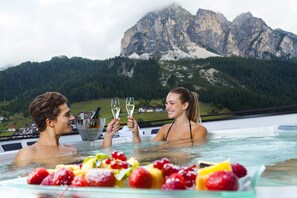 A couple in a hot tub with a mountain view, holding glasses of champagne, and a fruit bowl in the foreground.