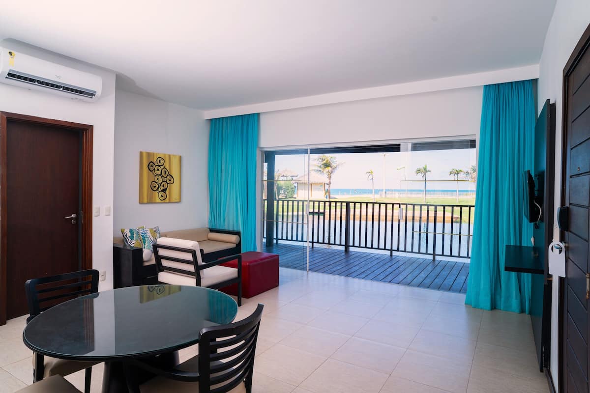A modern room with a glass table and chairs, a sofa, and a red ottoman. It features a balcony with a view of the sea and palm trees.