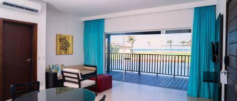 A modern room with a glass table and chairs, a sofa, and a red ottoman. It features a balcony with a view of the sea and palm trees.