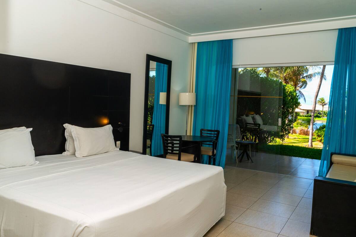 A hotel room with a large bed, a desk, and a chair. There is a window with blue curtains and a view of a garden.