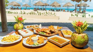 A table with various dishes and drinks, a coconut, and a view of a beach with umbrellas and lounge chairs. - Dom Pedro Laguna Beach Resort & Golf by WAM Experience (Aquiraz)