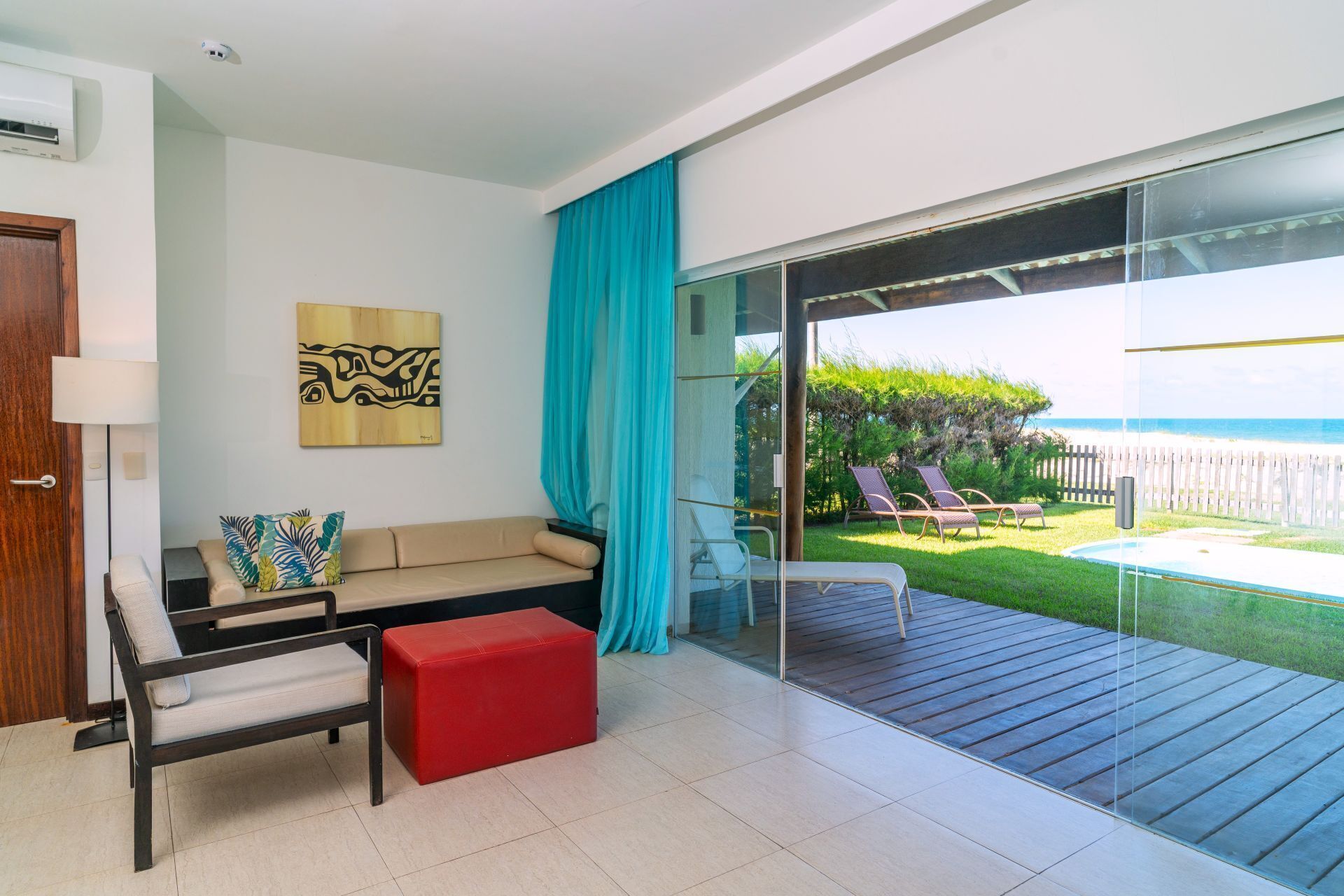 Un soggiorno moderno con un divano, un poggiapiedi rosso e una porta vetrata che dà su un balcone con vista sull'oceano e una piscina.