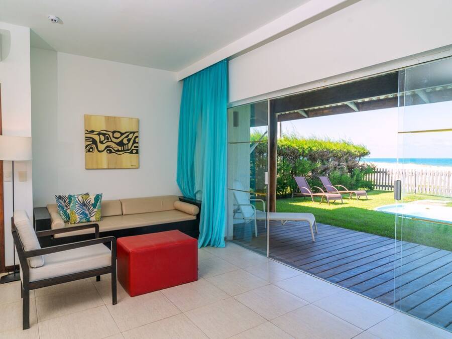 Un soggiorno moderno con un divano, un poggiapiedi rosso e una porta vetrata che dà su un balcone con vista sull'oceano e una piscina.