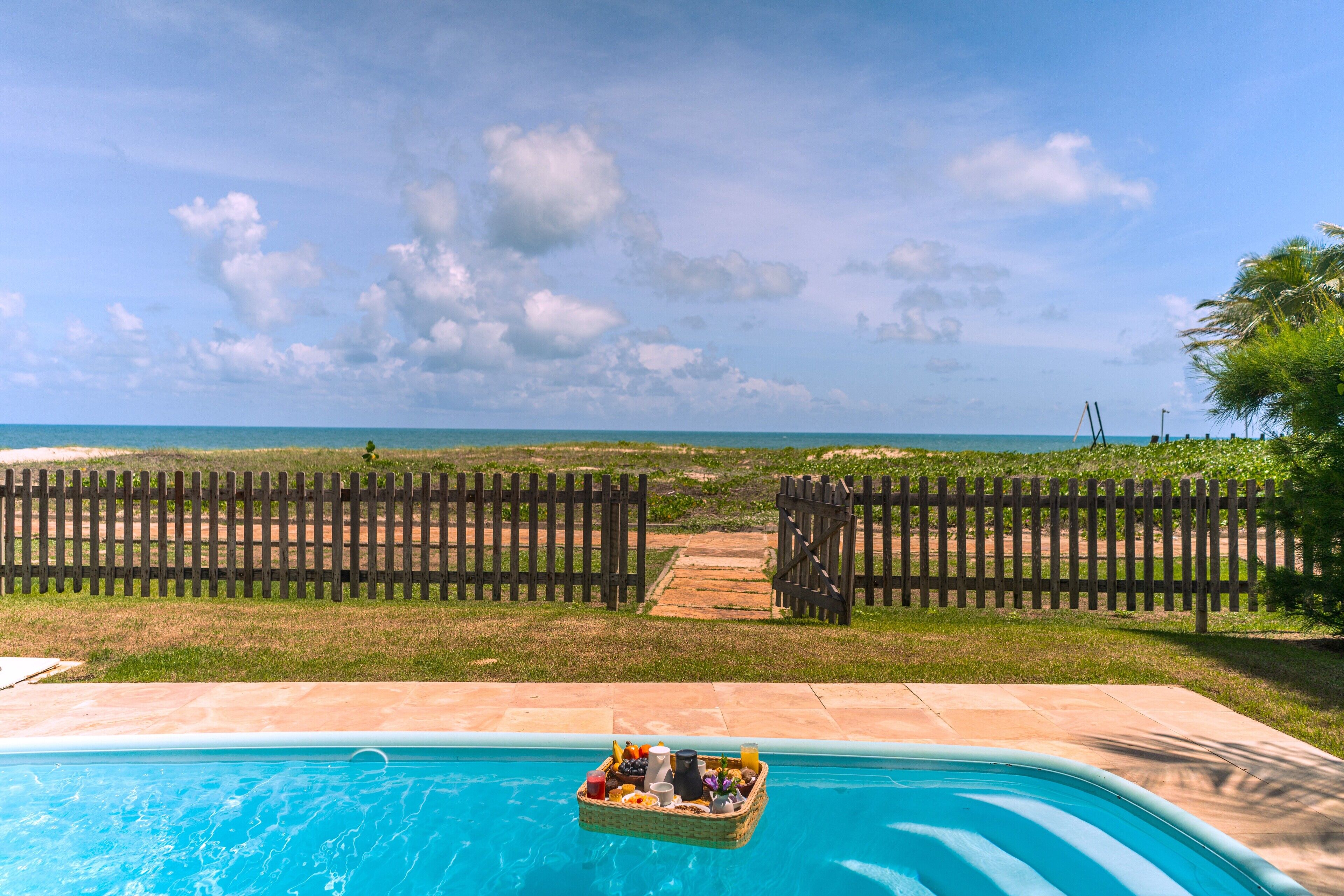 Una piscina con una cesta flotante de comida y bebidas, rodeada por una valla de madera y vista al océano.