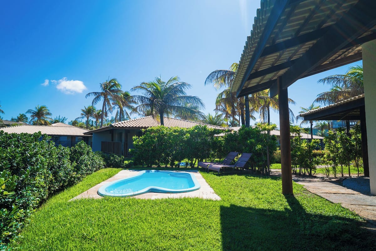 A swimming pool surrounded by lush greenery and palm trees, with a covered patio area and a building with a tiled roof in the background.