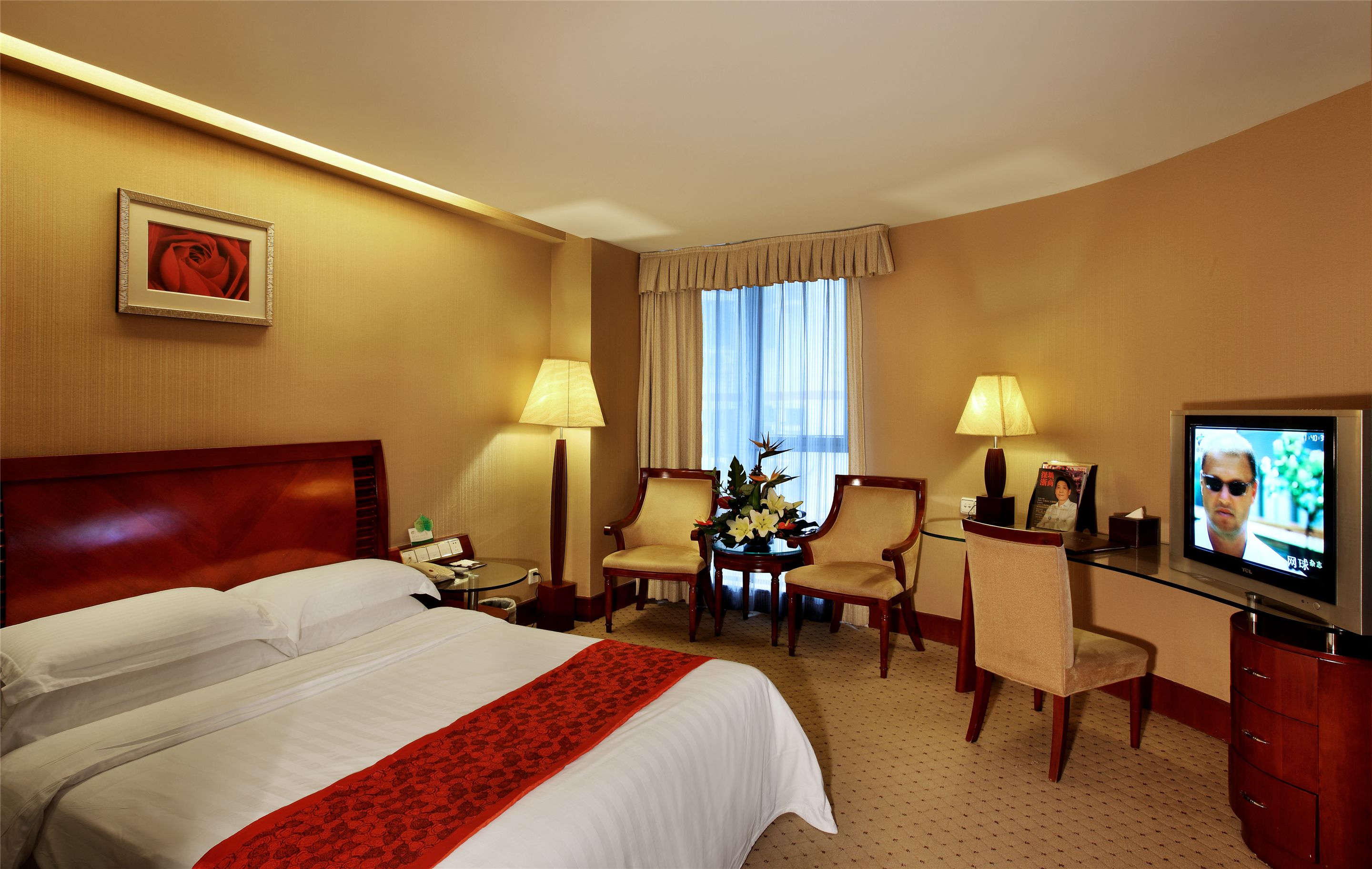 Quarto de hotel com uma cama grande, uma mesa com televisão, duas cadeiras, uma pequena mesa com flores e um quadro na parede.