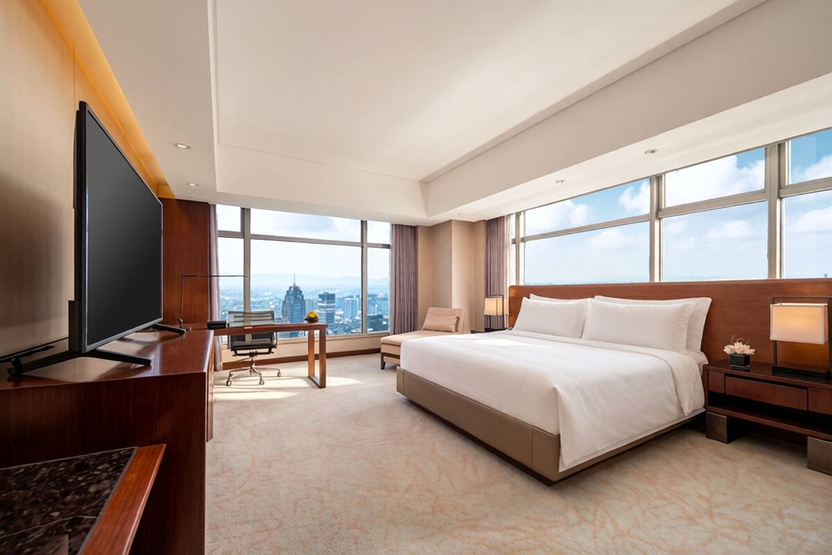Een hotelkamer met een groot bed, een bureau, een stoel, een televisie en grote ramen met uitzicht op de stad.