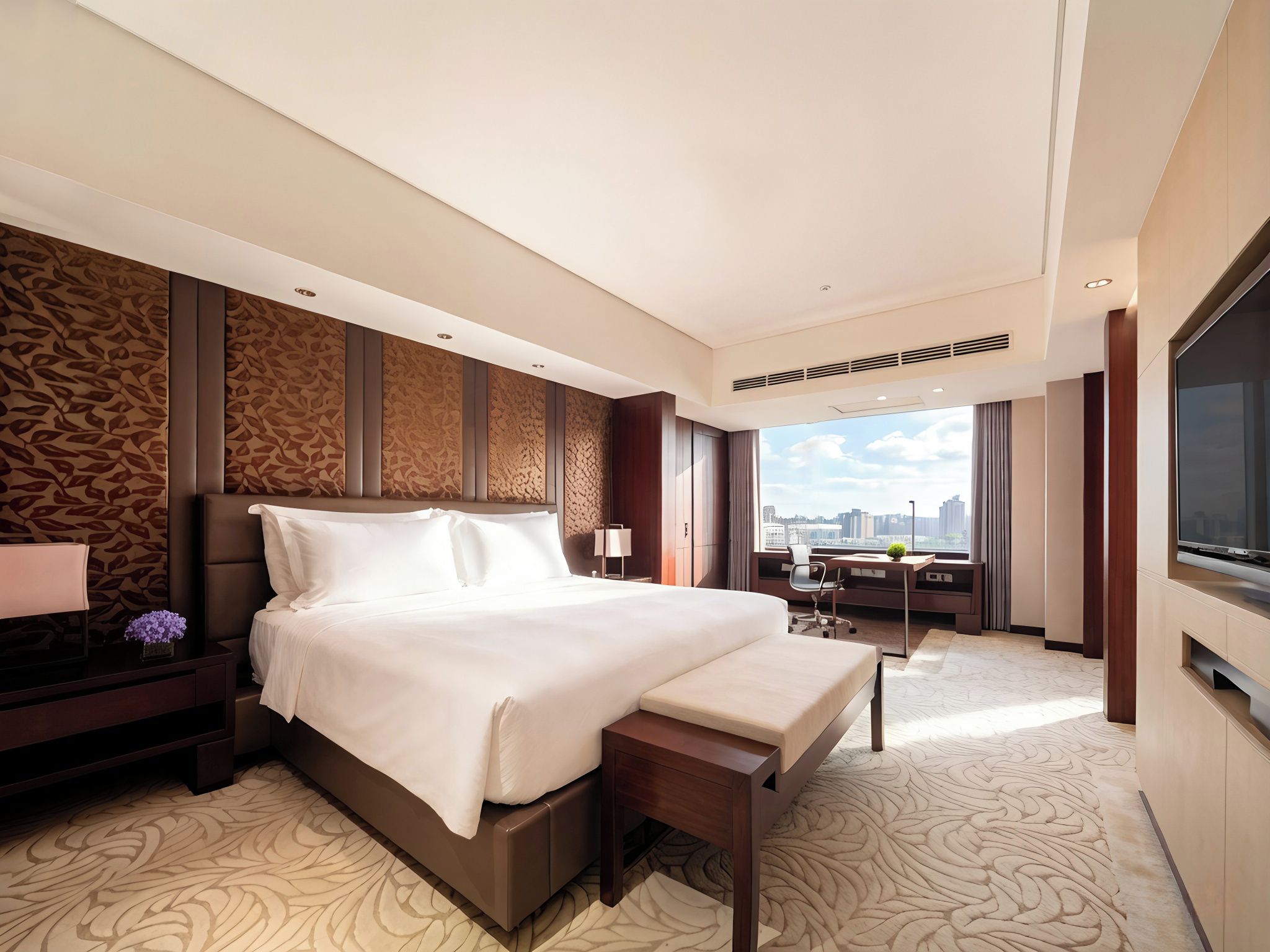 A modern hotel room with a large bed, a desk, a chair, a TV, and a city view.