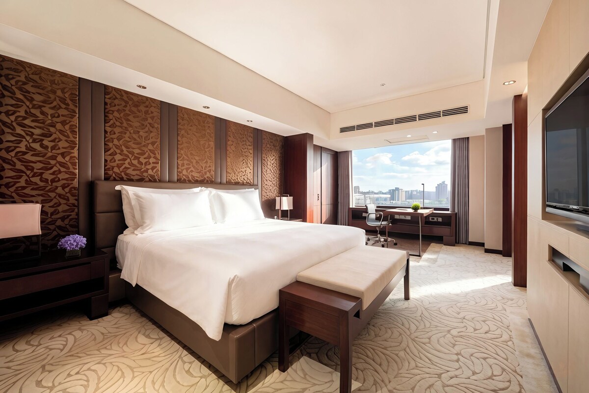 A modern hotel room with a large bed, a desk, a chair, a TV, and a city view.