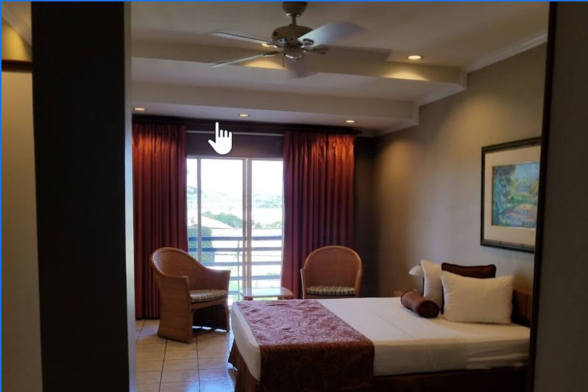 A hotel room with a bed, two chairs, a ceiling fan, and a large window with curtains.