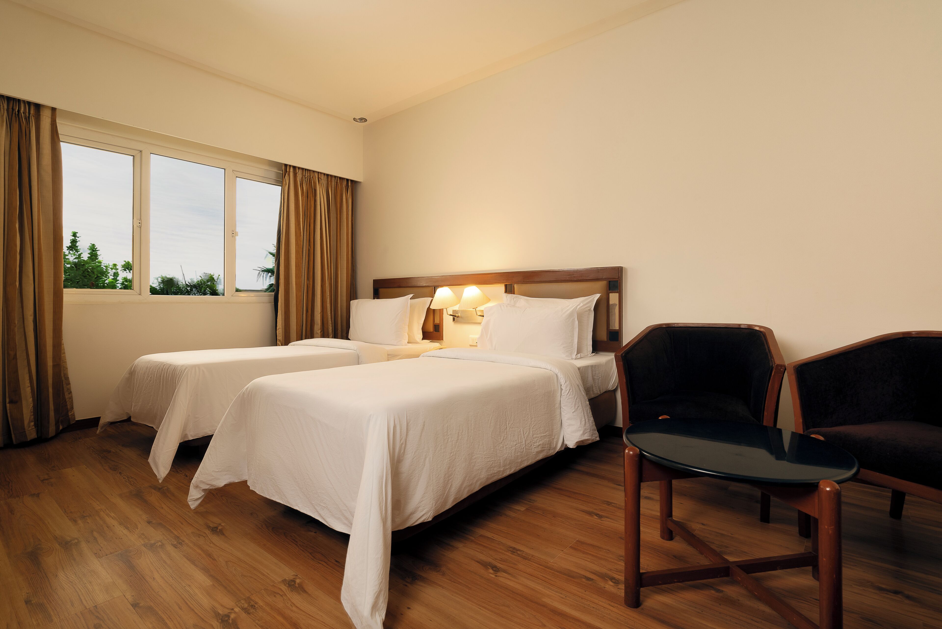 A hotel room with two beds, a wooden floor, a window with curtains, and a table with chairs.