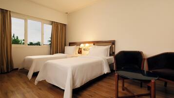 A hotel room with two beds, a wooden floor, a window with curtains, and a table with chairs.