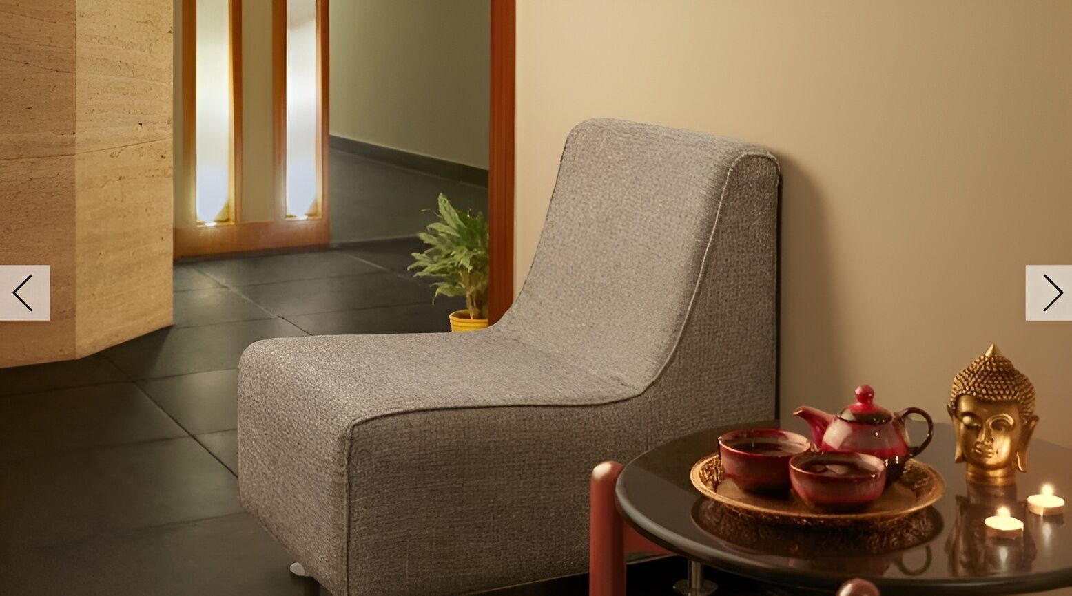 A modern room with a grey upholstered armchair, a small table with a teapot and cups, and a Buddha statue.