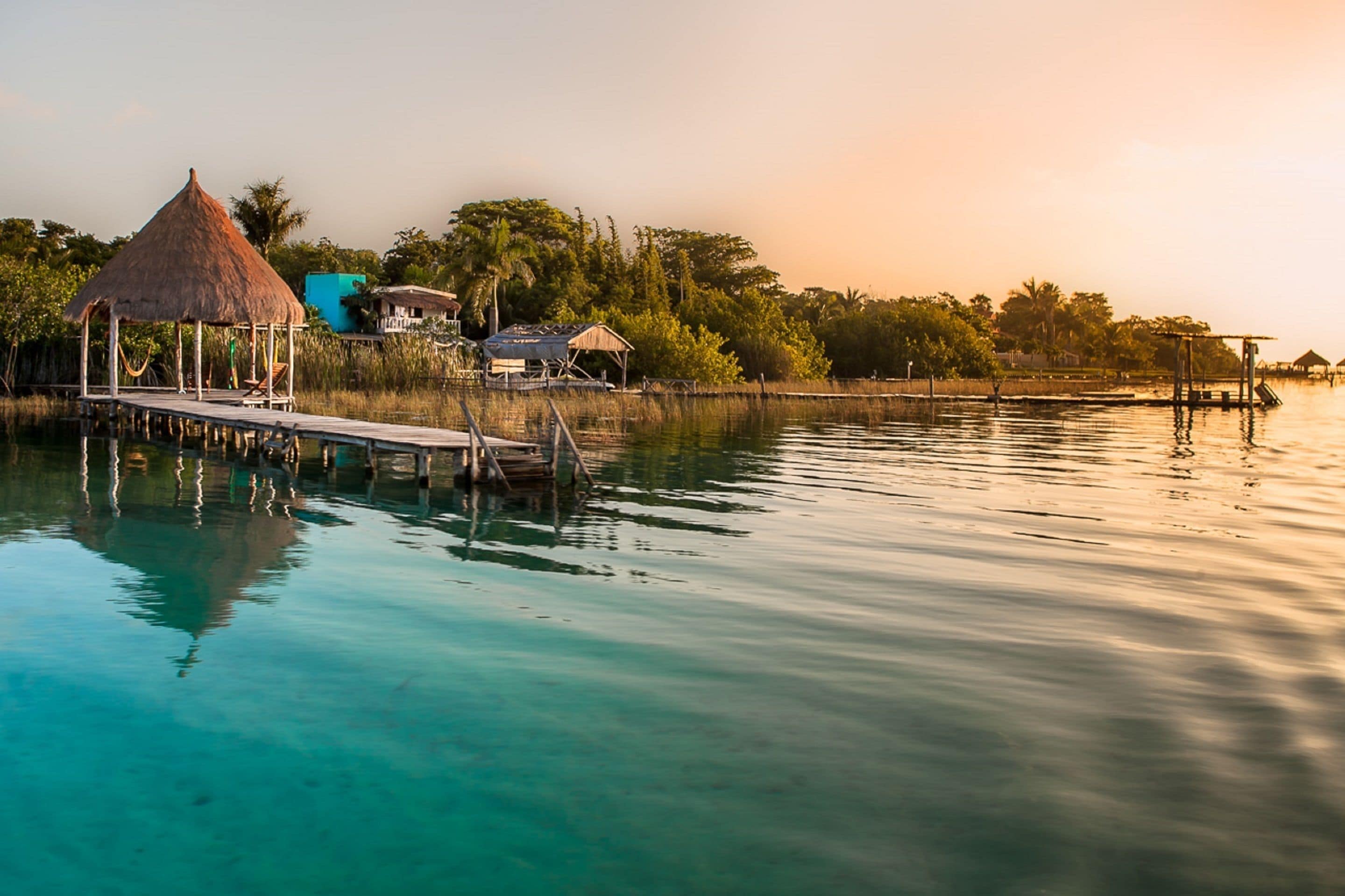 A calm waterfront with a thatched-roof structure, a wooden dock, and several buildings in the background.