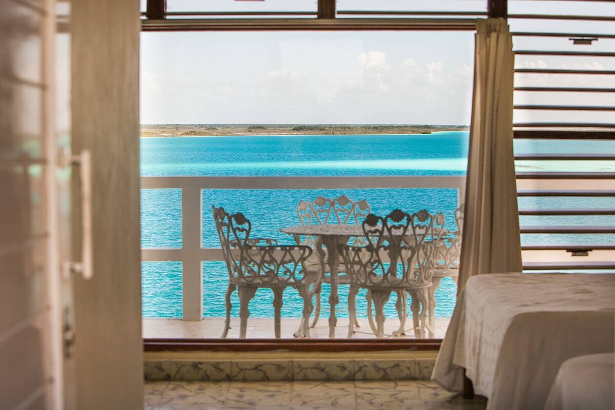 A room with a view of a turquoise sea and a balcony with white metal chairs and a table.
