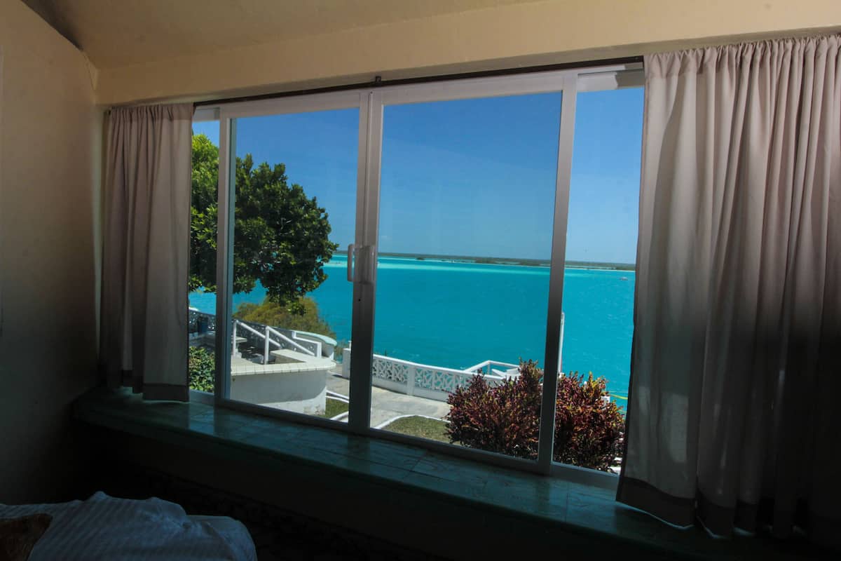 A room with a large window overlooking a clear blue sea and a white building with a staircase.