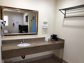 A bathroom with a granite countertop, a sink, a mirror, a hairdryer, a towel rack, and a coffee maker.