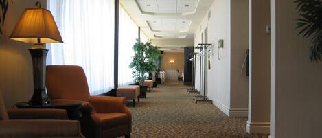 A hotel lobby with a carpeted floor, a seating area with chairs, a lamp, a plant, and a hallway leading to a door.