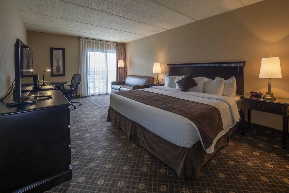 A hotel room with a large bed, two bedside tables with lamps, a desk with a computer, a chair, and a view of the city through the window.