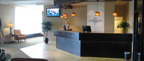 A hotel reception area with a reception desk, chairs, a television, and a potted plant.