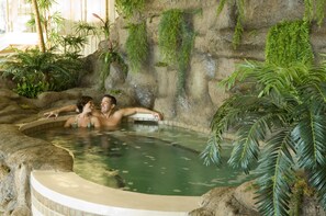 A hot tub with two people, surrounded by tropical plants and a rocky wall. - Hôtel Le Président (Sherbrooke)