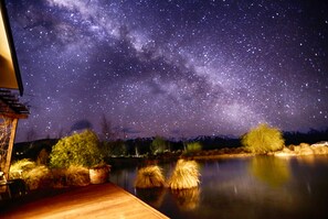 A night scene with a starry sky, a wooden deck, and a calm body of water reflecting the stars. - Matuka Luxury Lodge (Ben Ohau)